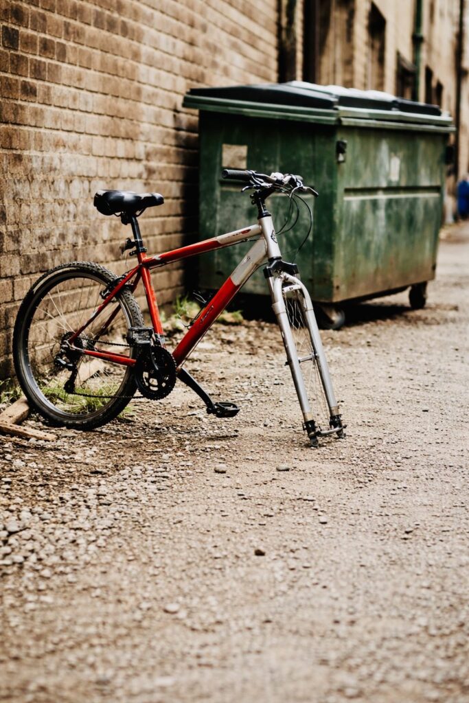 Ten-speed bicycle missing a wheel in a gravel alley next to a dumpster