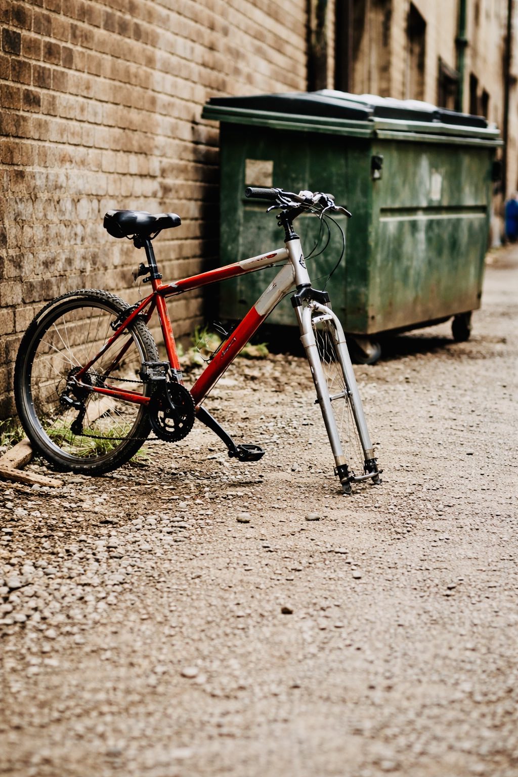 Ten-speed bicycle missing a wheel in a gravel alley next to a dumpster